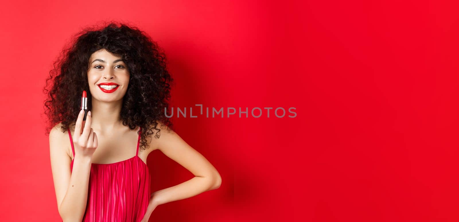 Fashionable woman with curly hair, showing red lipstick and smiling, recommend cosmetic, standing on white background by Benzoix