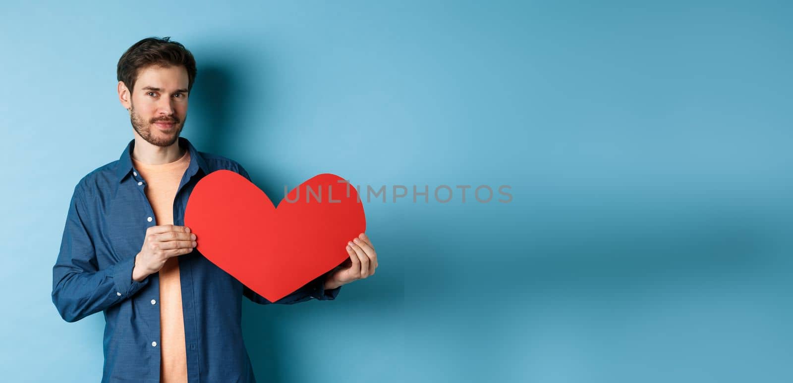 Handsome bearded guy showing valentines day cutout and smiling, standing in casual clothes over blue background. Copy space