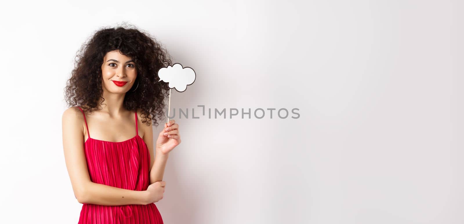 Elegant woman with curly hair, wearing red evening dress, holding cloud and smiling, standing on white background.