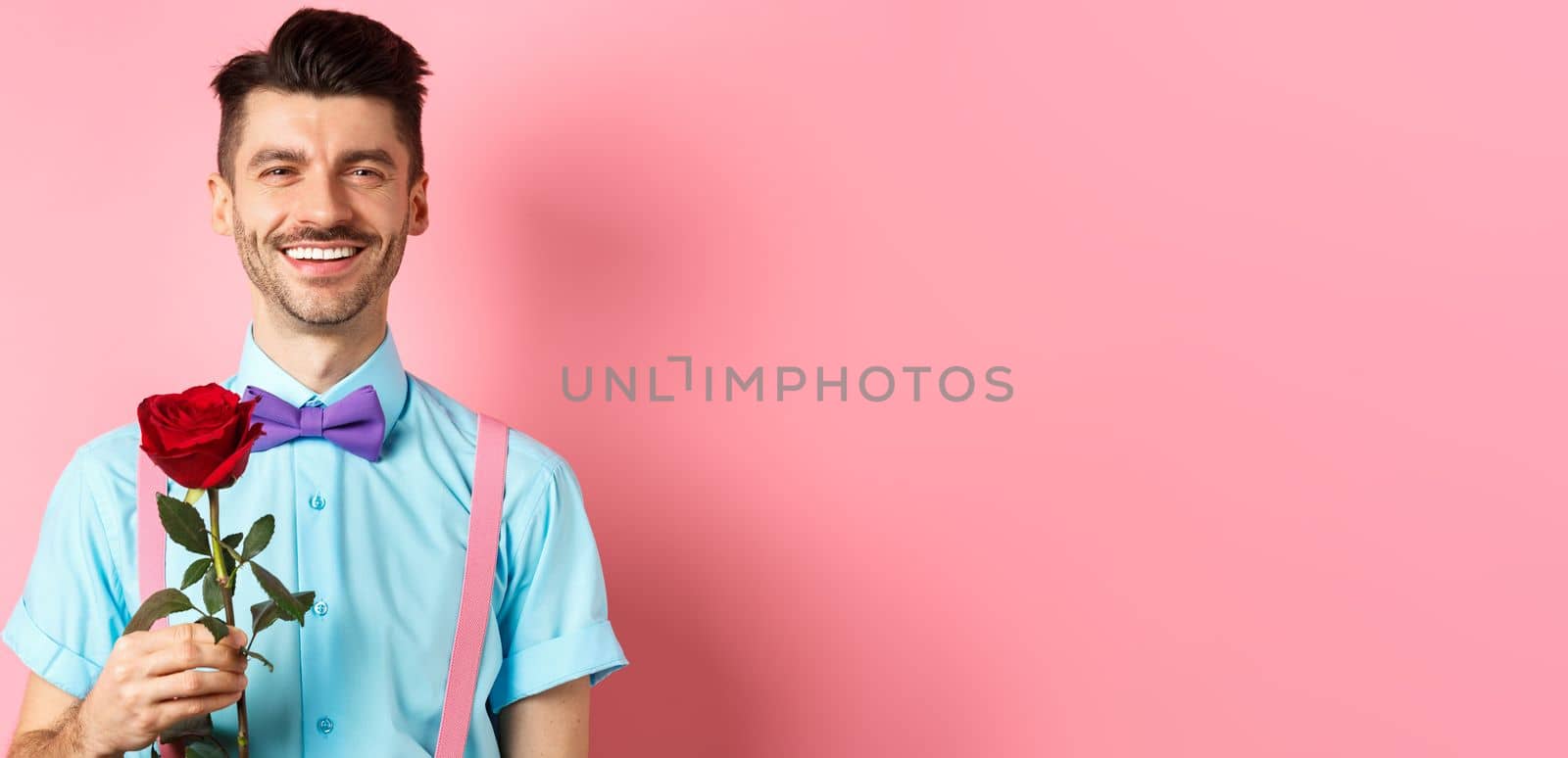 Attractive fancy guy waiting for date on lovers date, holding red rose and smiling, standing on romantic pink background. Valentines day concept.