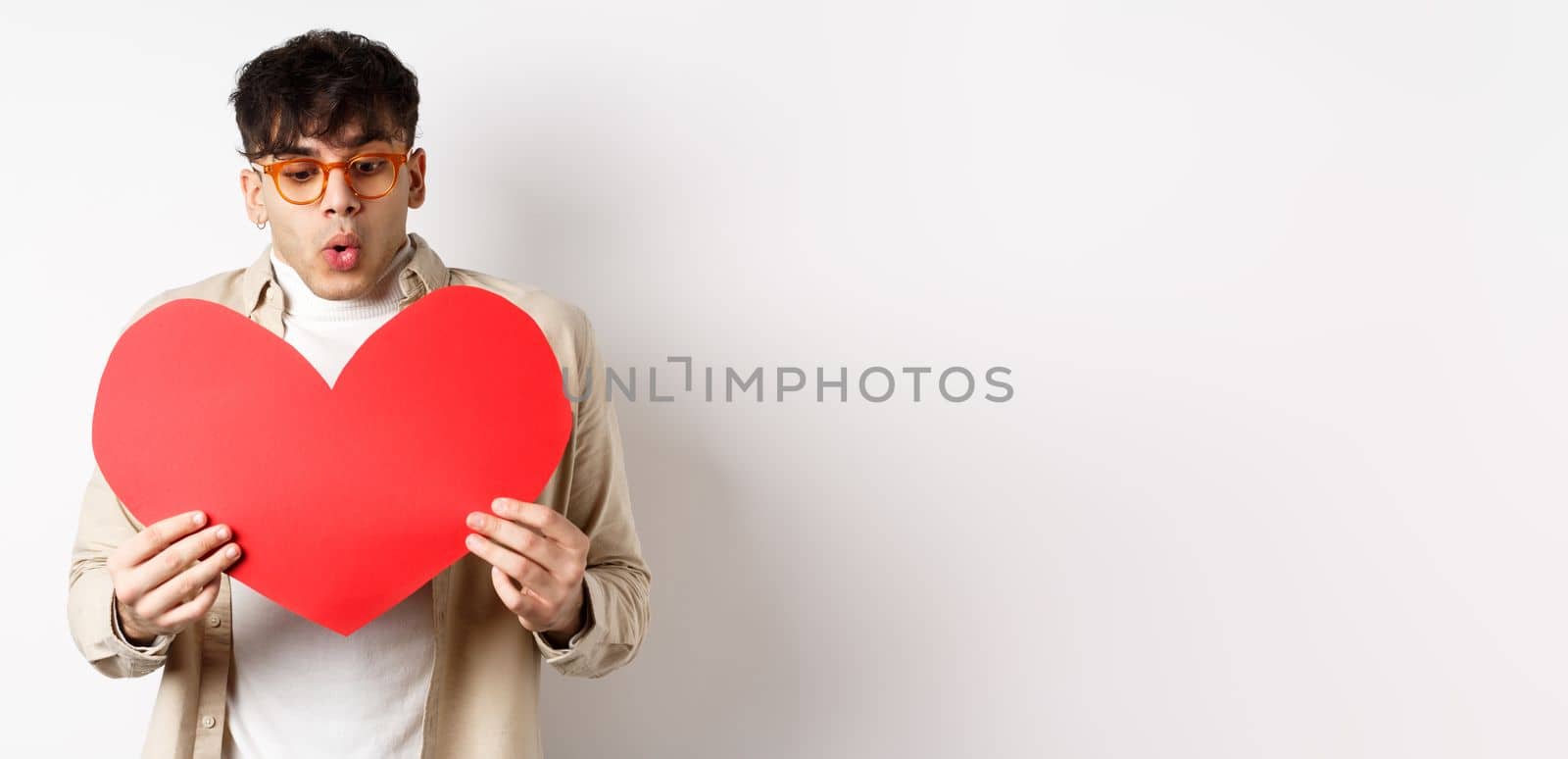 Surprised handsome man receive big red heart postcard on Valentines day, looking at gift with amazement, enjoying lovers day, standing over white background by Benzoix