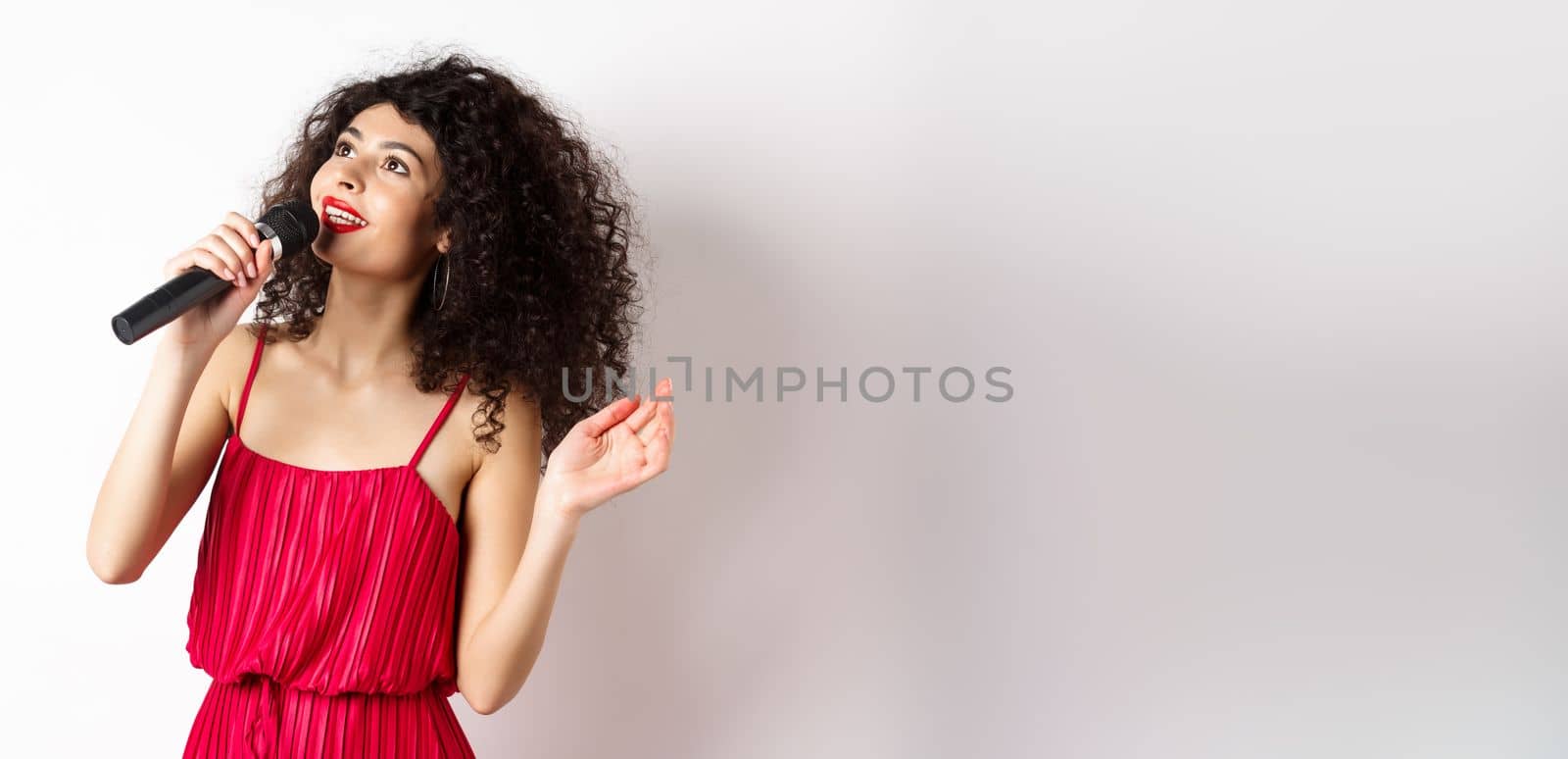 Beautiful lady in red dress singing songs in microphone, smiling and looking up, standing on white background.