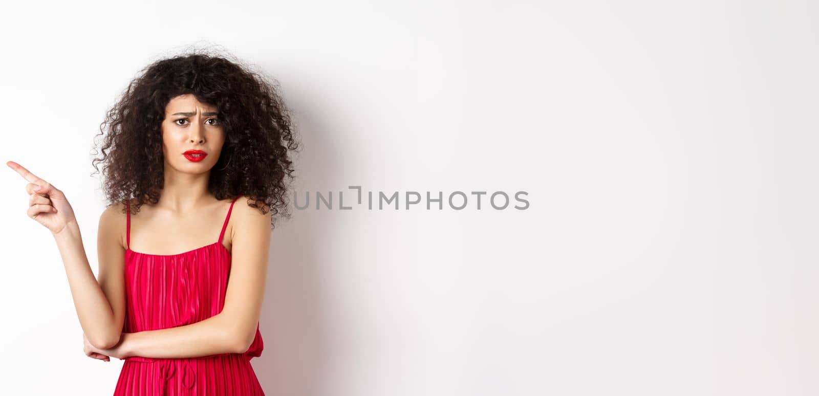 Arrogant young woman with curly hair, wearing red dress, frowning and complaining, pointing finger left at promo, standing over white background.