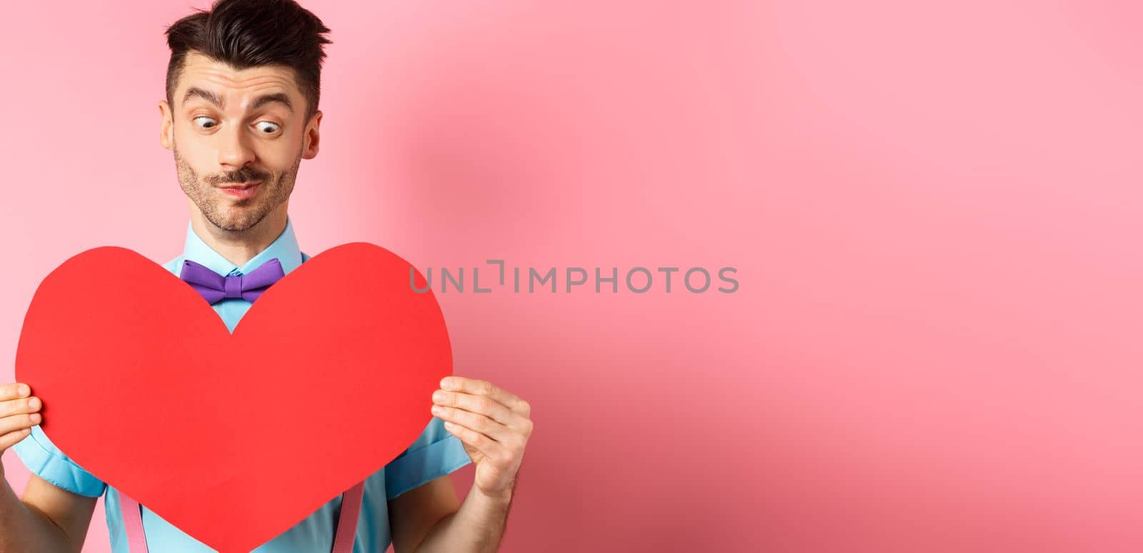 Valentines day concept. Cute boyfriend looking at big red heart, waiting for lover on romantic date, pink background by Benzoix