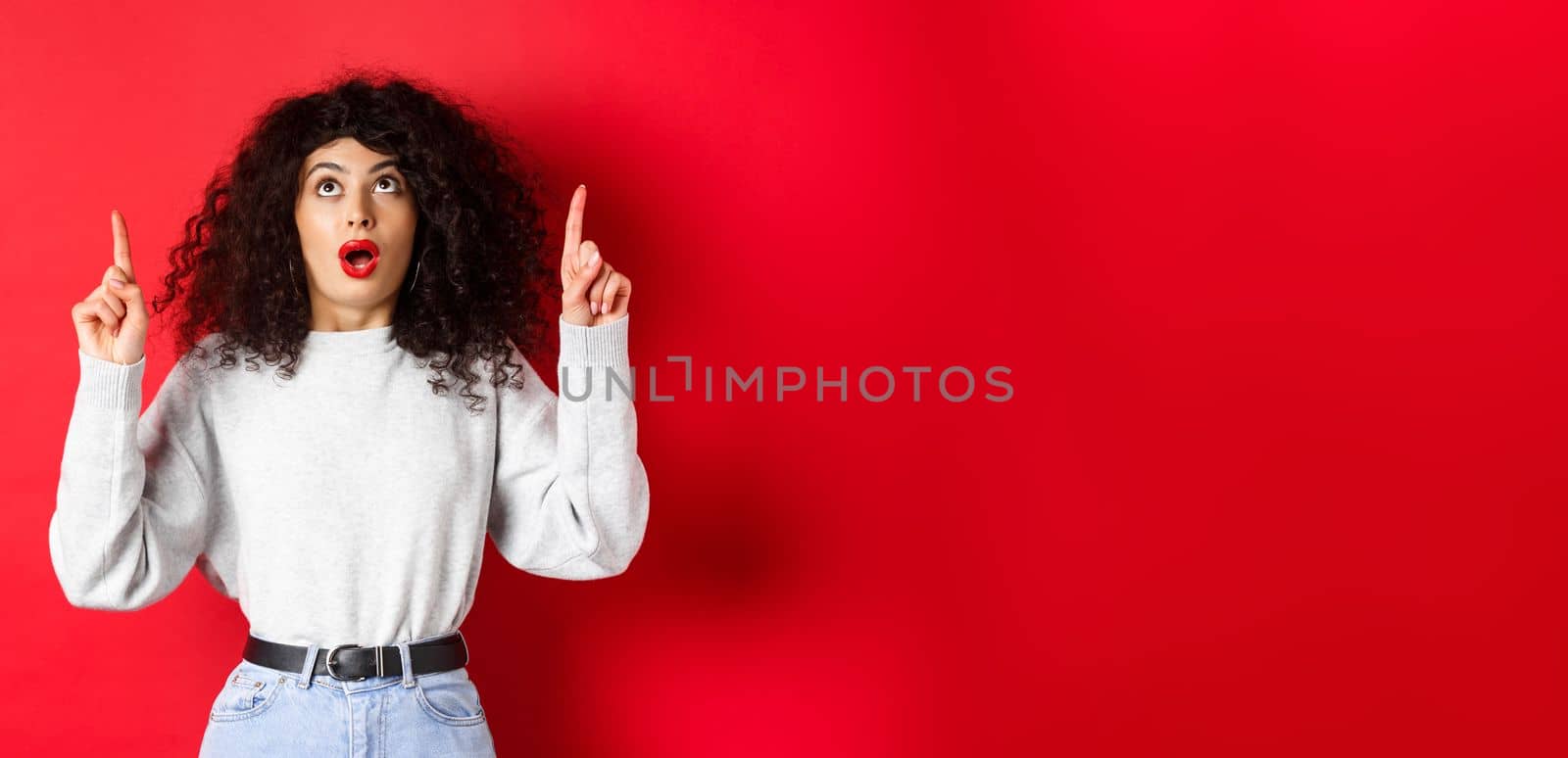 Impressed european female model in casual clothes, checking out promo with dropped jaw, looking and pointing fingers up, showing logo, standing on red background.