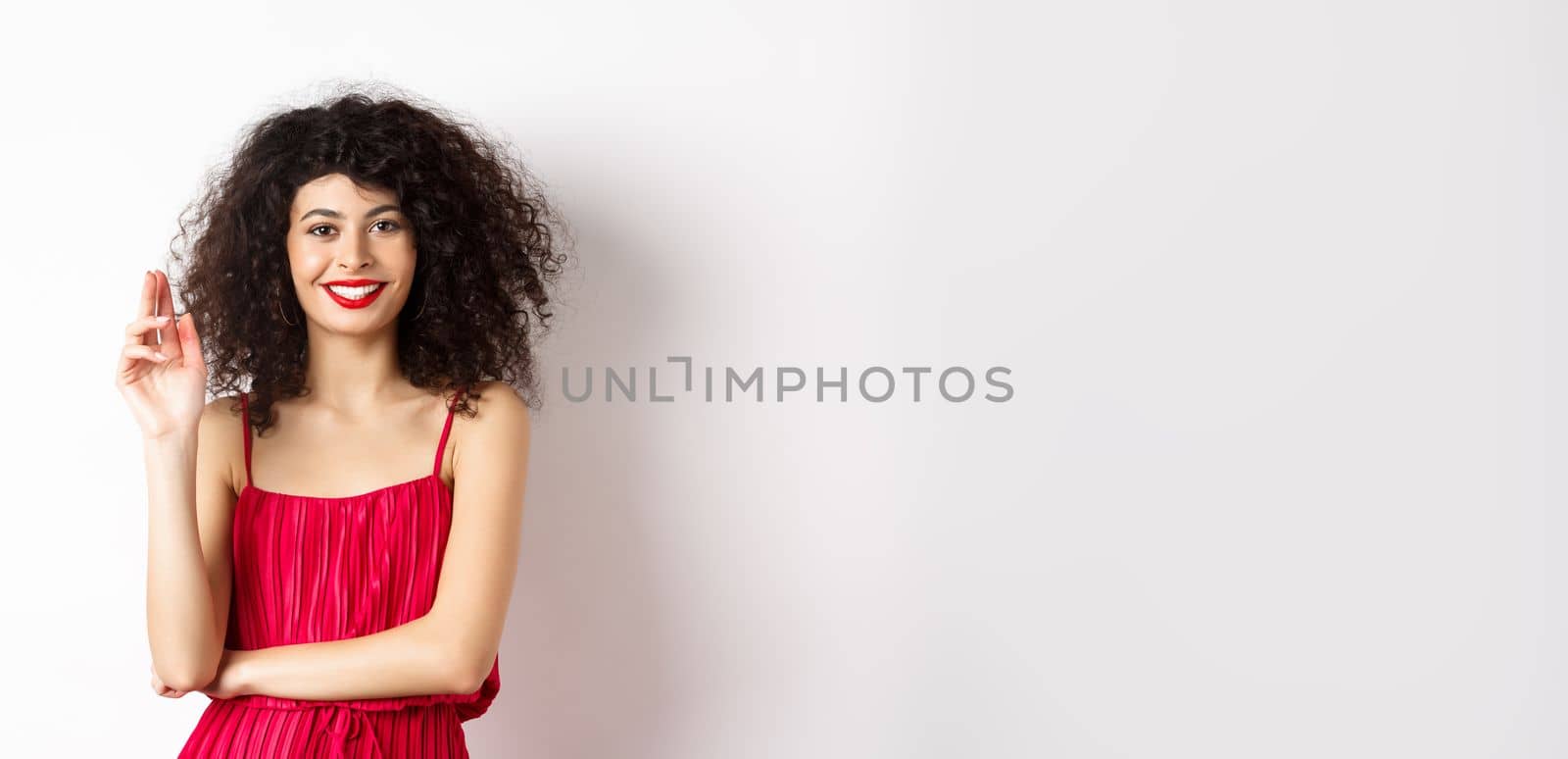 Beauty and fashion. Smiling woman with curly hair and makeup, wearing red dress, waving hand in greeting gesture, saying hello, standing on white background.