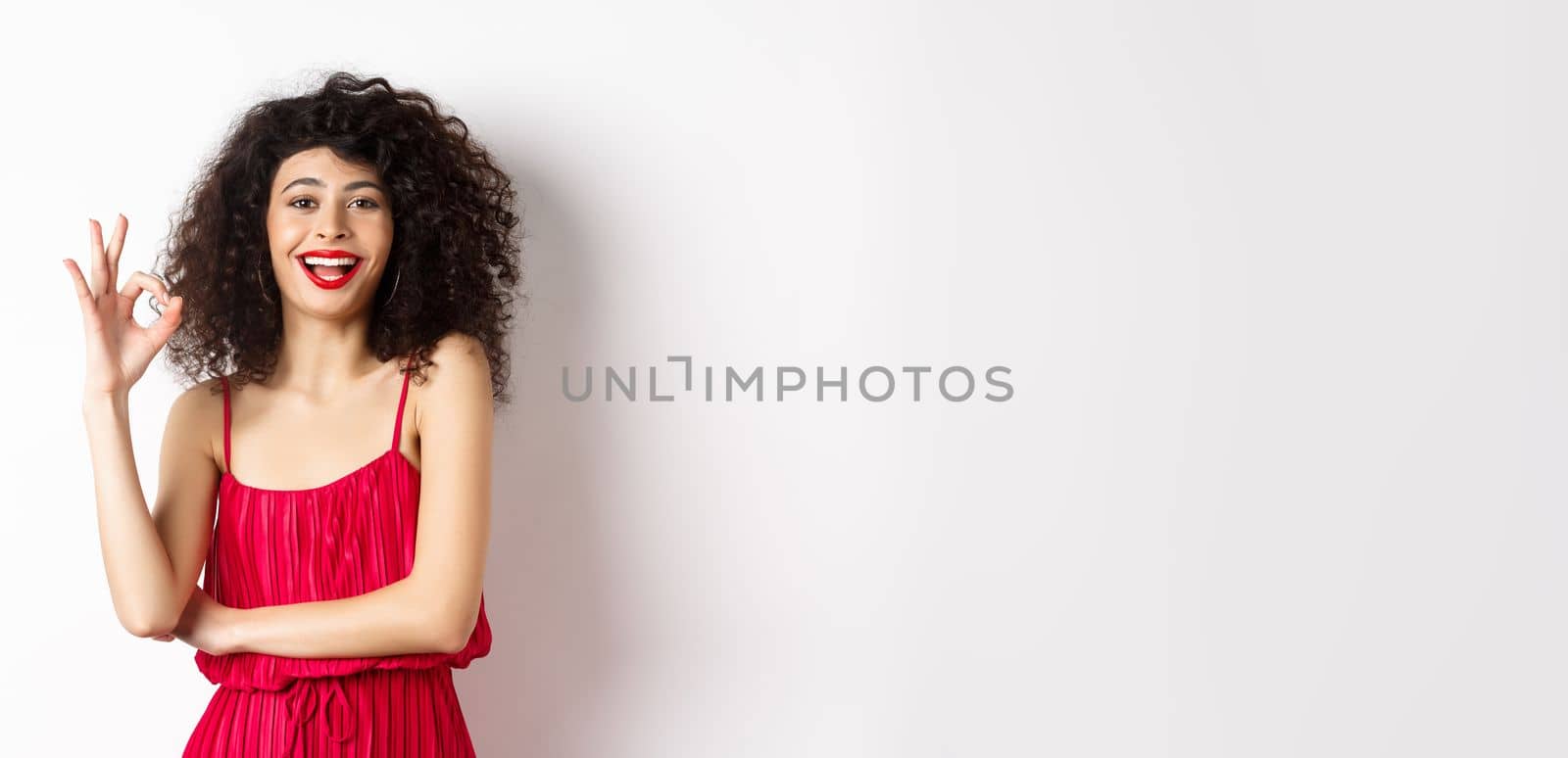 Cheerful caucasian woman with curly hair and makeup, wearing elegant red dress, showing OK sign and smiling in approval, standing over white background by Benzoix