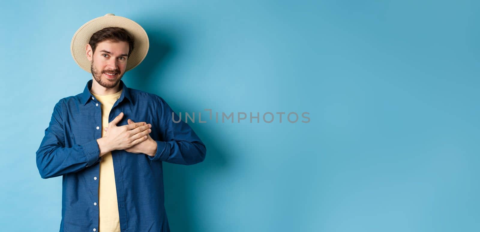 Handsome traveler in straw hat, holding hands on heart and smiling, showing thank you gesture, keep memories about vacation in soul, standing on blue background.