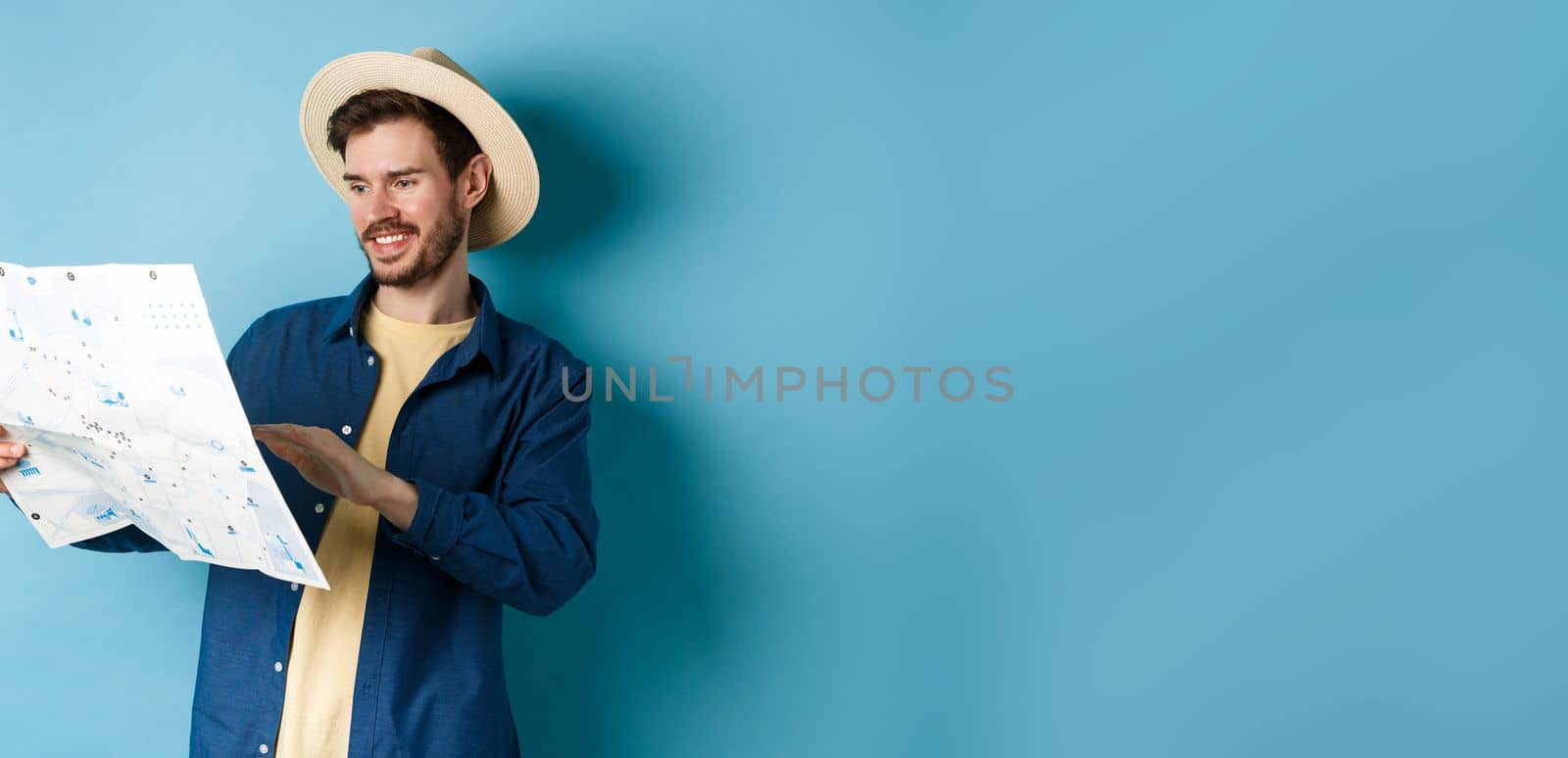 Handsome smiling tourist in straw hat looking at map, choosing travel road, planning a vacation, standing on blue background.