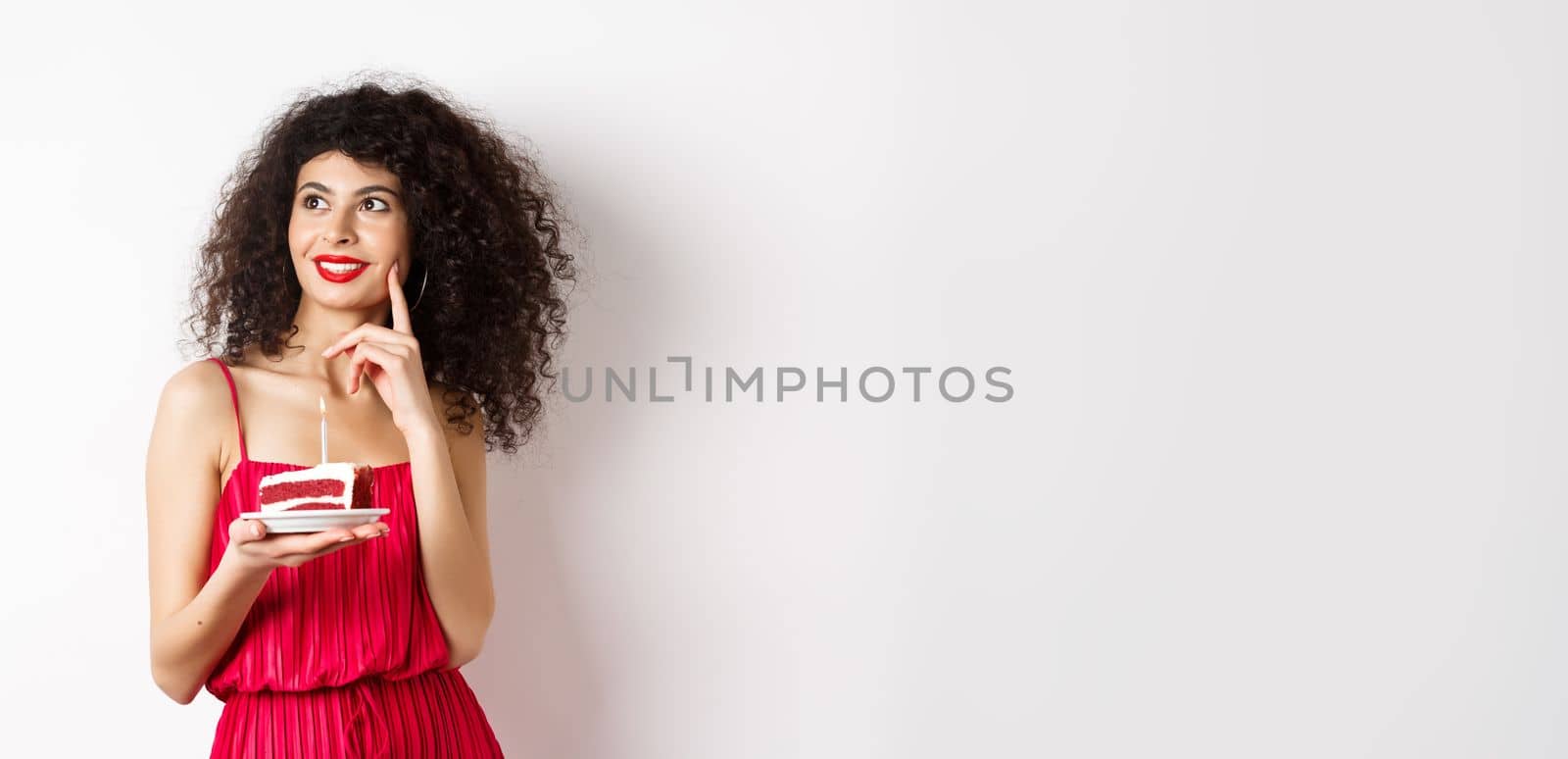 Happy woman celebrating birthday, holding cake and making wish, looking aside dreamy, standing in red dress on white background. Celebration and holiday concept by Benzoix