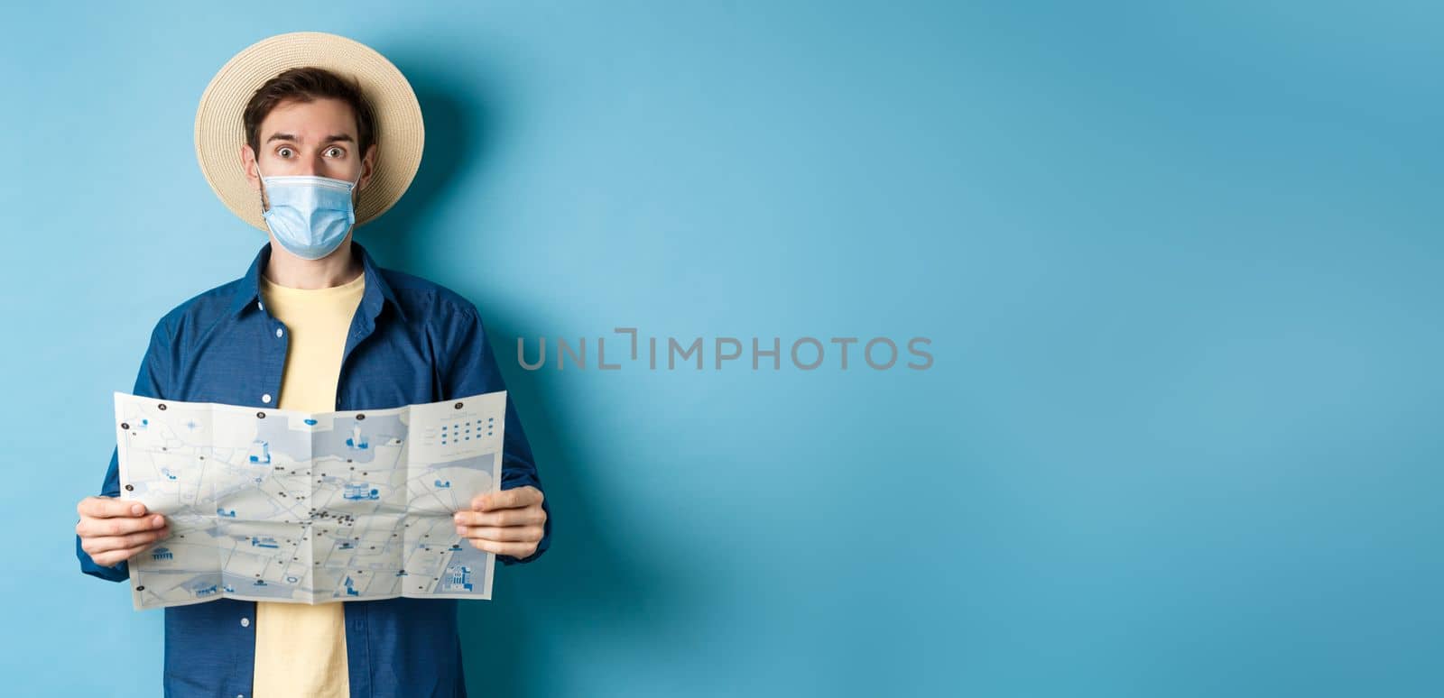 Covid-19, pandemic and travel concept. Tourist in medical mask look surprised, holding map, standing on blue background.