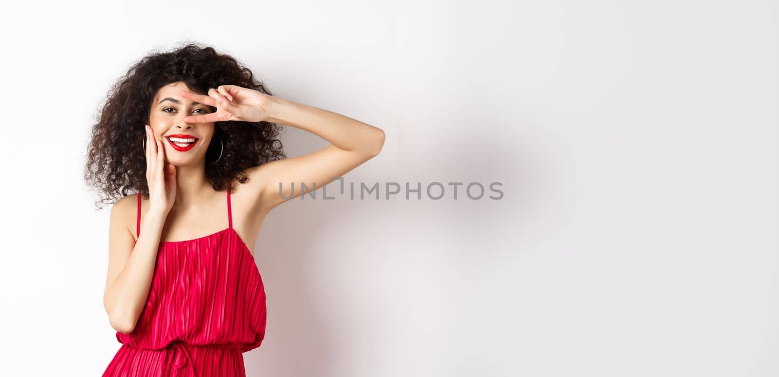 Beauty and fashion. Romantic woman with curly hair and red dress, showing v-sign and smiling happy at camera, standing on white background by Benzoix