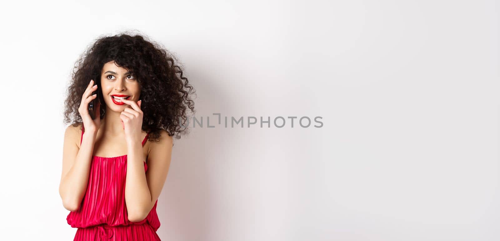 Silly fashionable woman in red dress and lips, biting fingernail duing phone call, thinking on mobile conversation, standing on white background.