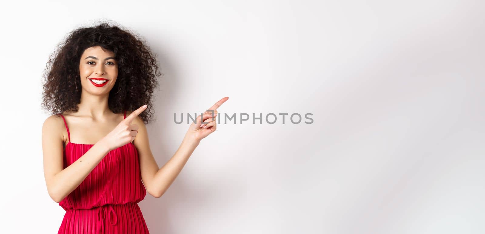 Cheerful female model in fashionable red dress, smiling and pointing fingers right at logo, standing over white background.