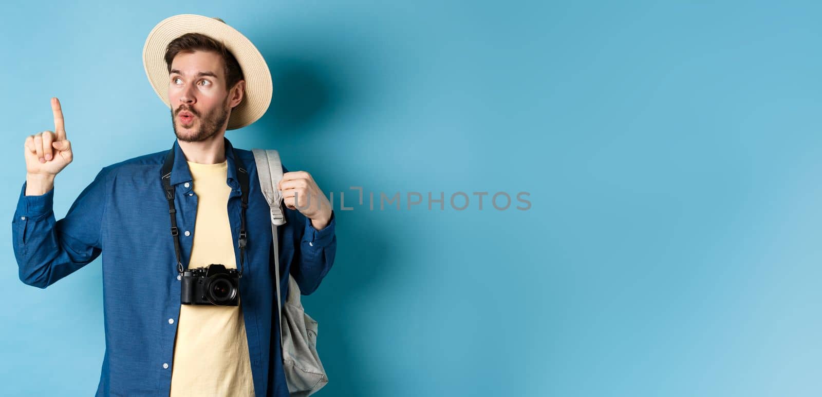 Handsome tourist looking curious and pointing up, showing interesting sightseeing on summer vacation, wearing straw hat and holding backpack with camera, blue background.