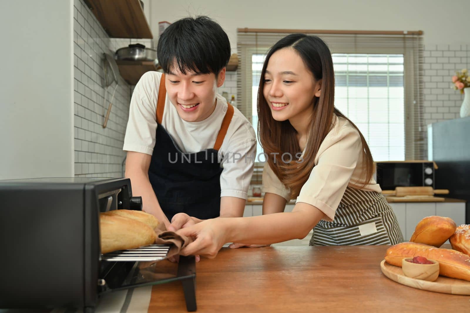 Exited married couple taking off french baguette bread from oven, preparing dinner in modern kitchen together by prathanchorruangsak