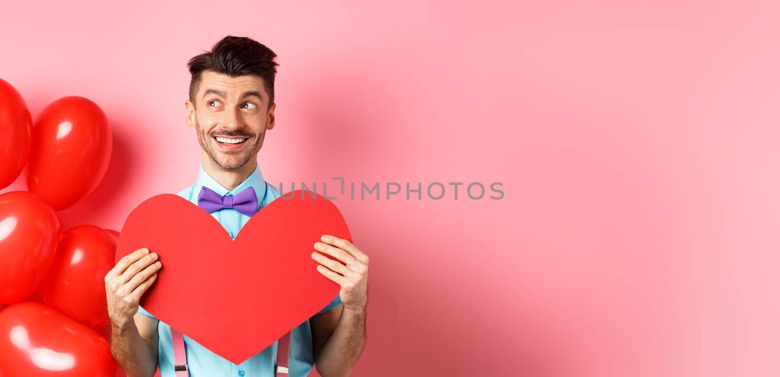 Valentines day concept. Romantic guy smiling and looking left, dreaming of date with lover, showing red big heart cutout, pink background.