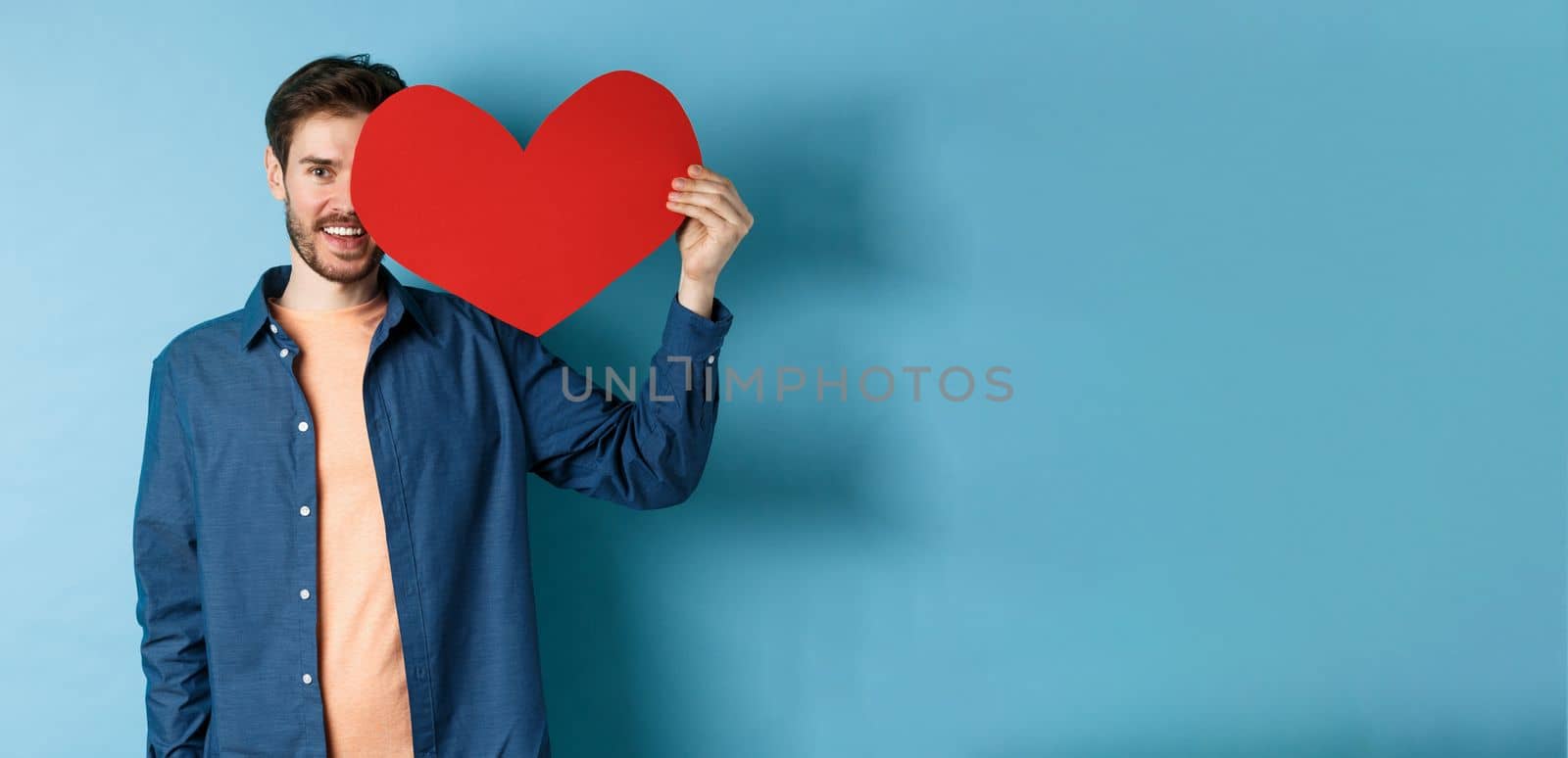 Happy man showing valentines heart and smiling, make romantic gift on lovers day, standing over blue background.