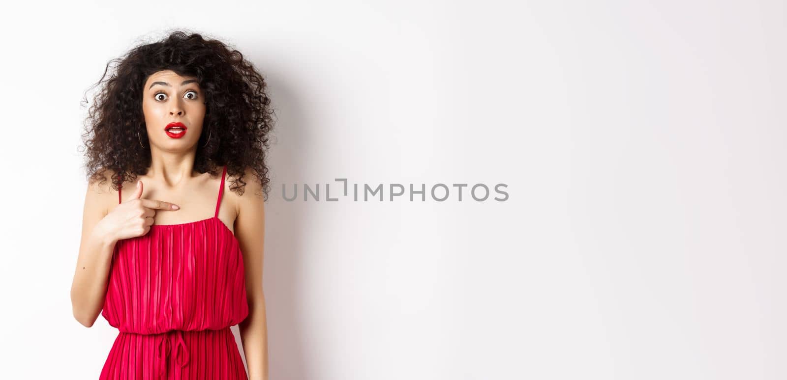 Surprised young woman in red dress pointing at herself, being confused and shocked, standing on white background by Benzoix