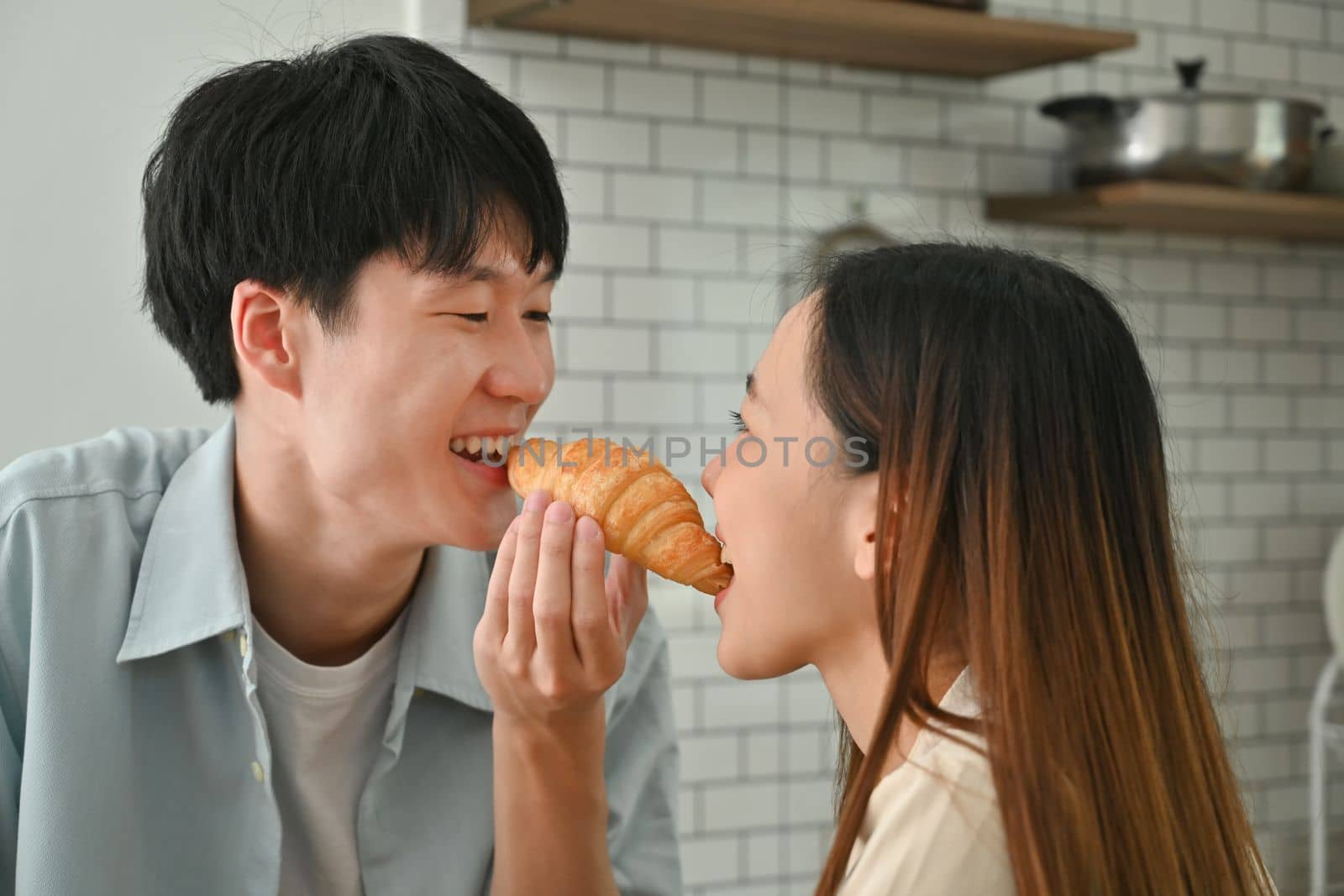 Cute young couple eating croissant while having breakfast in kitchen, enjoying leisure weekend time together by prathanchorruangsak