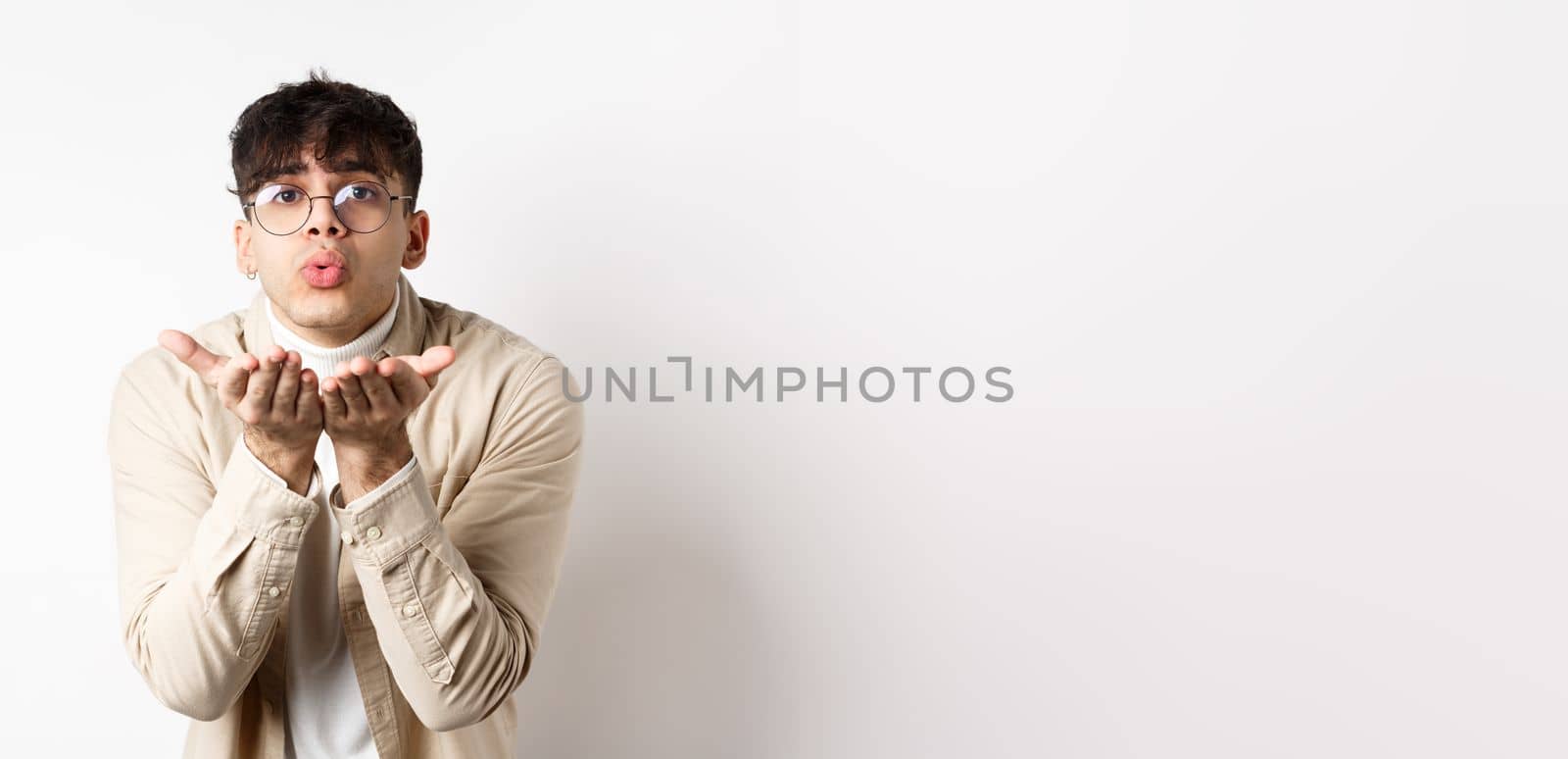 Cute boyfriend sending air kiss at camera on happy Valentines day, looking tender at lover, standing in casual clothes and glasses on white background.
