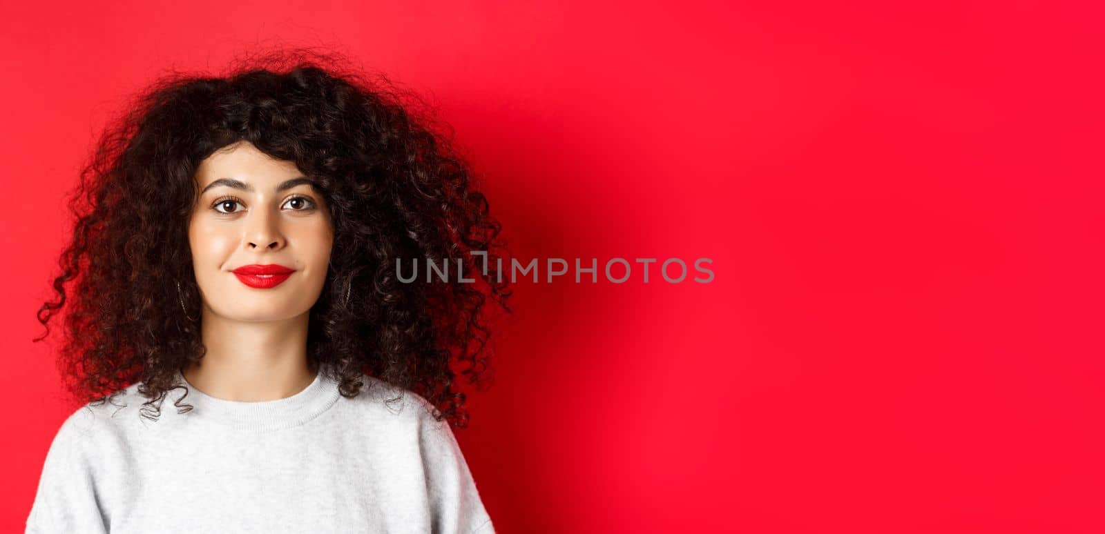 Close up portrait of beautiful modern woman with curly hairstyle and red lips, smiling happy at camera, standing on studio background.