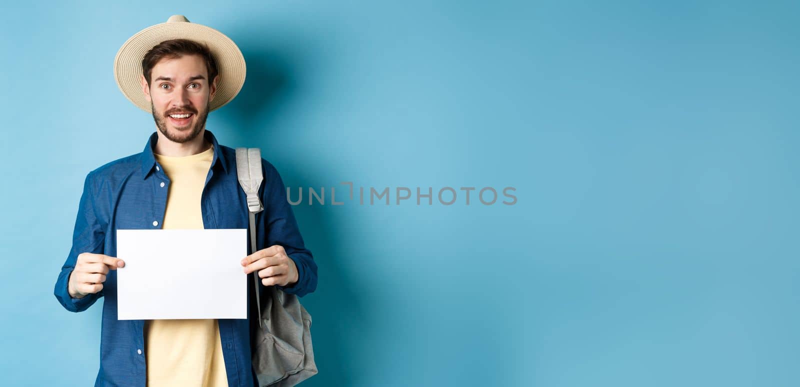 Cheerful tourist in summer hat, showing empty piece of paper and smiling, hitchhiking with backpack, standing on blue background.