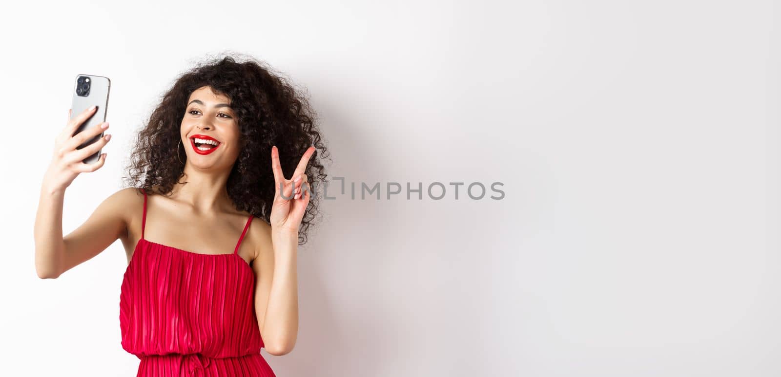 Elegant young woman in red dress taking selfie on smartphone, posing on event party, standing with mobile phone on white background.