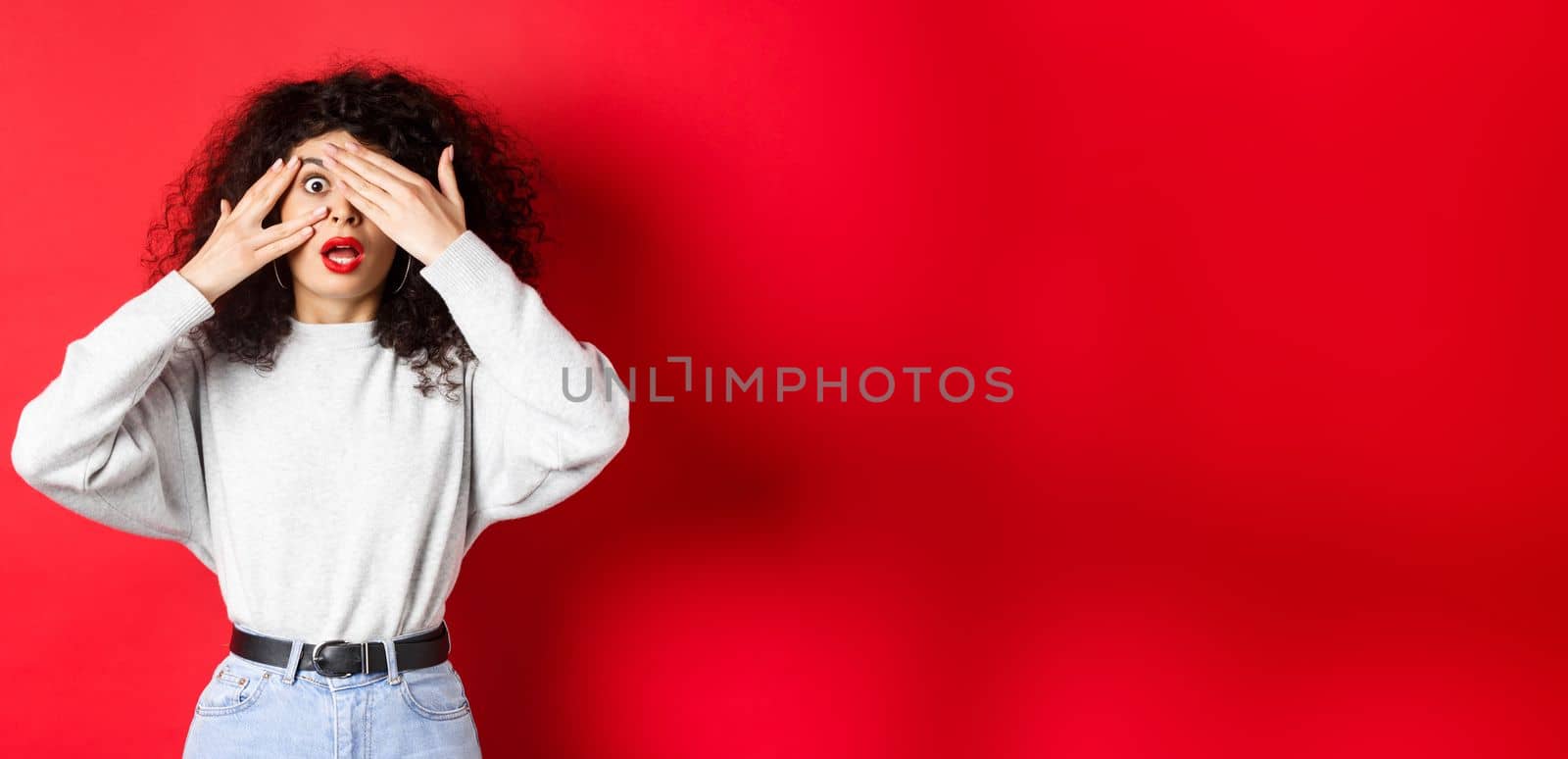 Shocked caucasian girl covering eyes and peek through fingers at something unbelievable, gasping speechless, standing on red background.