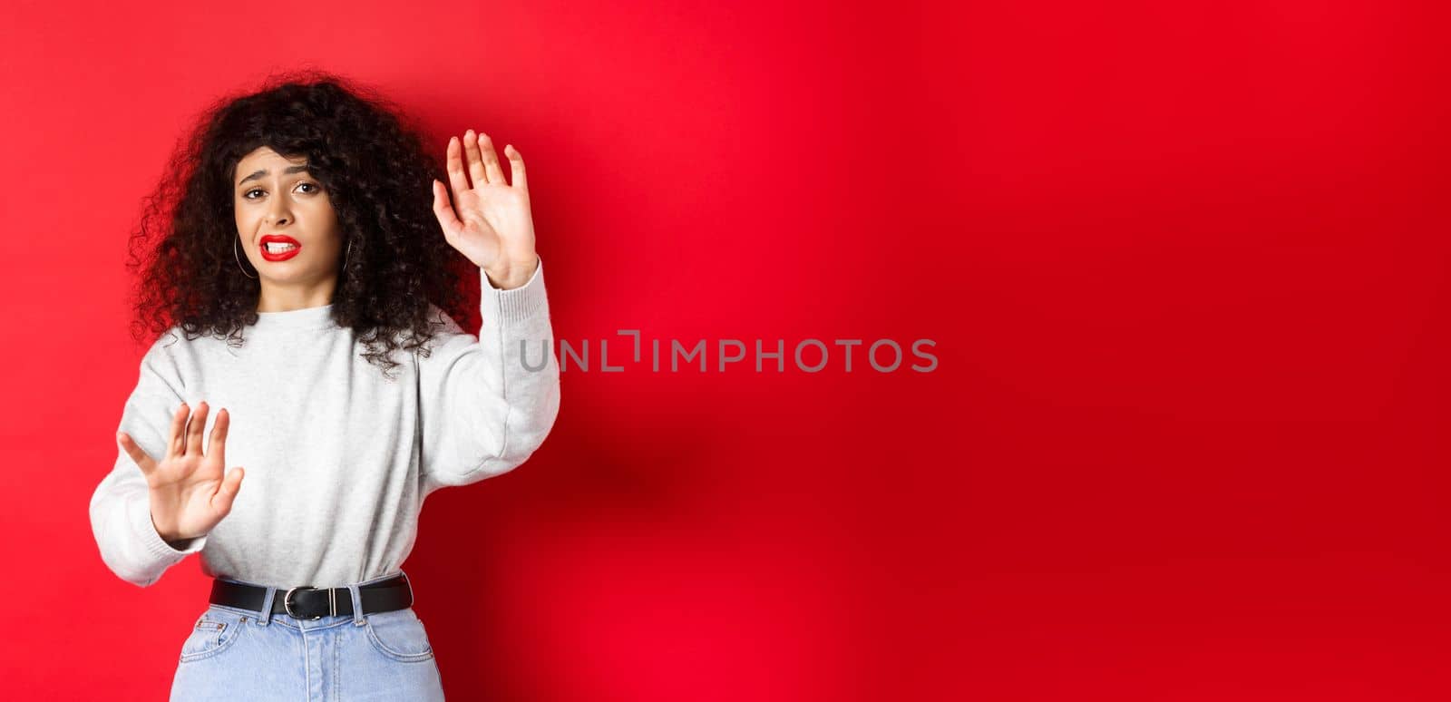 Disgusted young woman begging to stop, raising hands defensive, say no, standing on red background. Copy space