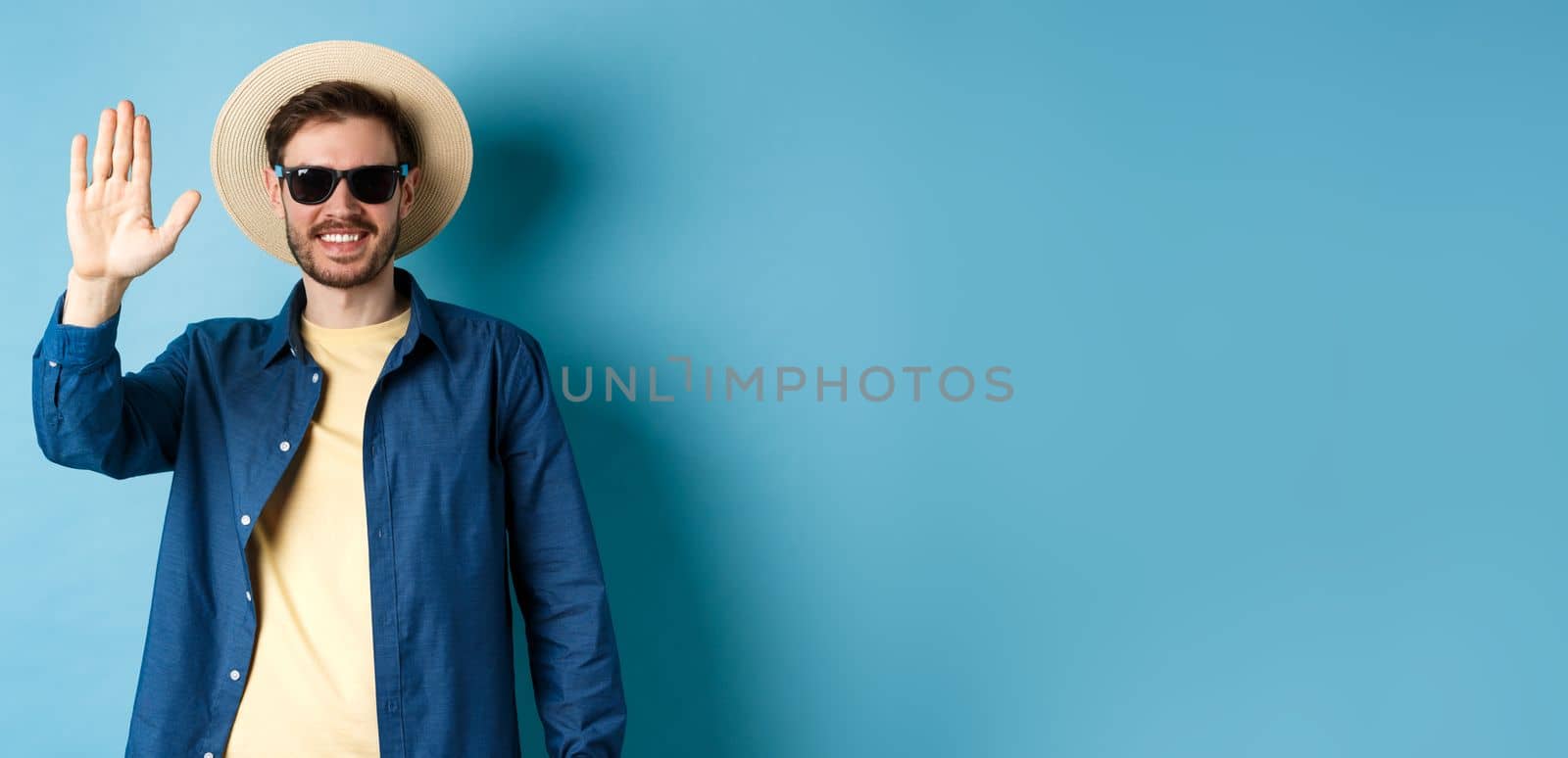 Handsome hipster on summer holiday, waiving hand and smiling, saying hello, wearing sunglasses and straw hat, blue background.