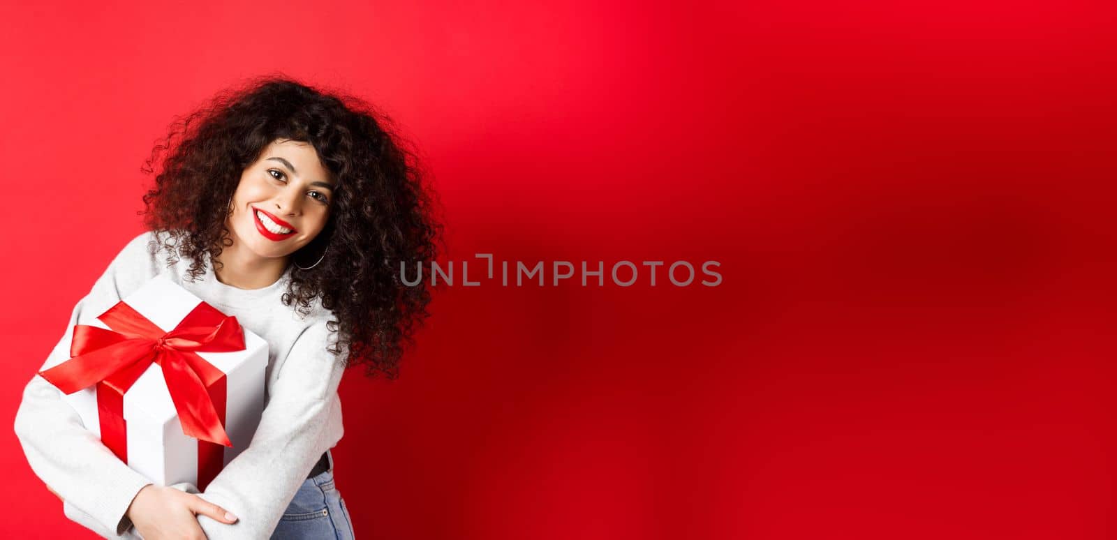 Celebration and holidays concept. Happy woman holding birthday gift and smiling at camera, standing in casual clothes, red background by Benzoix