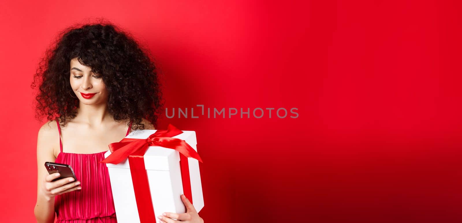 Valentines day and shopping. Beautiful caucasian woman in red dress, holding gift box from lover and using mobile phone, reading message on smartphone, studio background.