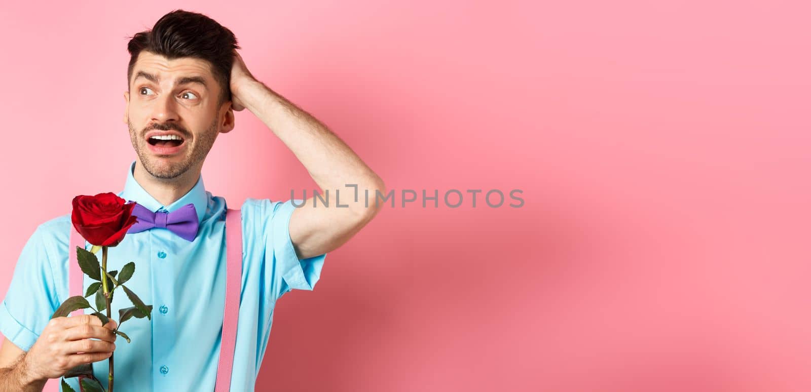 Nervous man waiting for his date on Valentines day, holding red rose and looking confused sideways, scratching head anxiously, standing on pink background.
