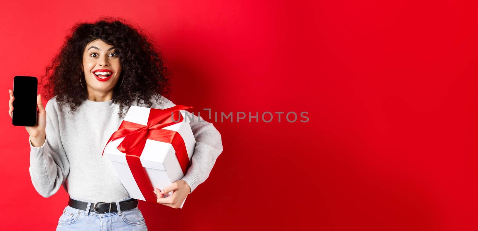 Valentines and lovers day. Excited smiling woman with curly dark hair, showing smartphone empty screen and holding surprise gift on holiday, showing online promo, red background by Benzoix