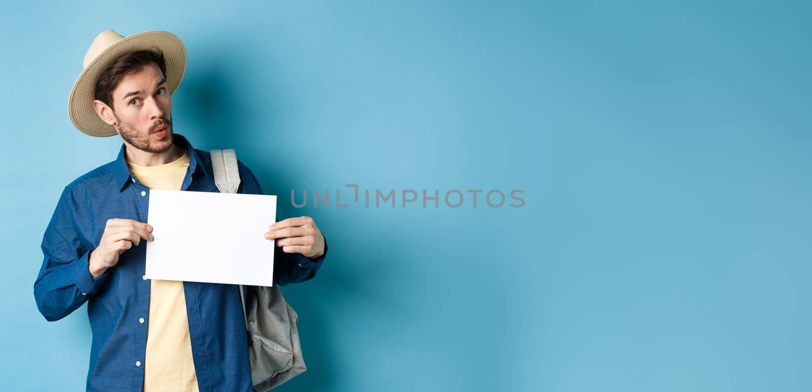 Excited tourist in straw hat, hitchhiking, showing blank piece of paper and looking amused, standing on blue background. Copy space