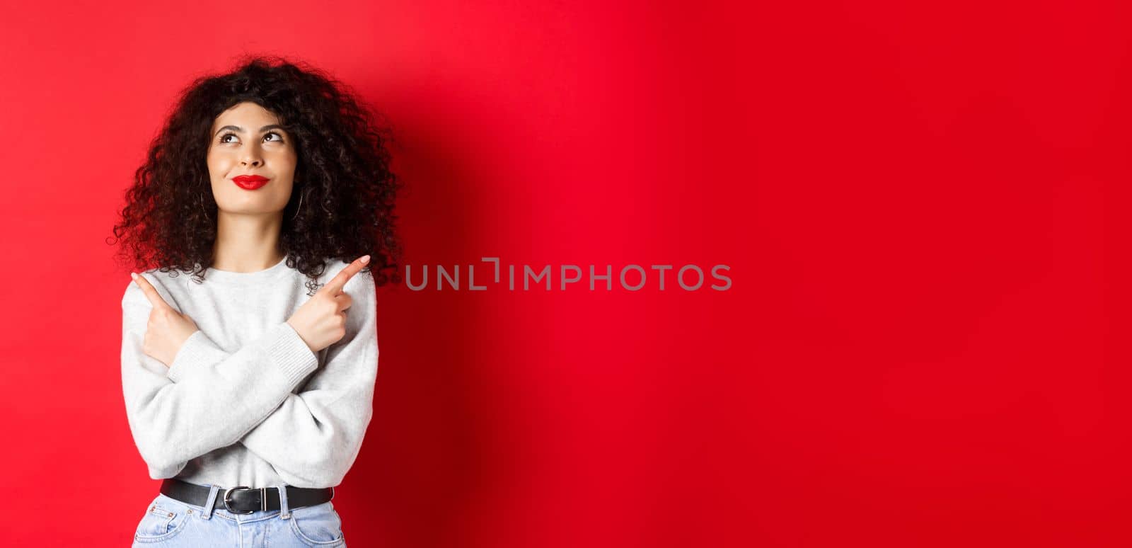 Image of happy smiling woman pointing sideways, looking up with determined face, choose something, standing confident in her decision on red background.