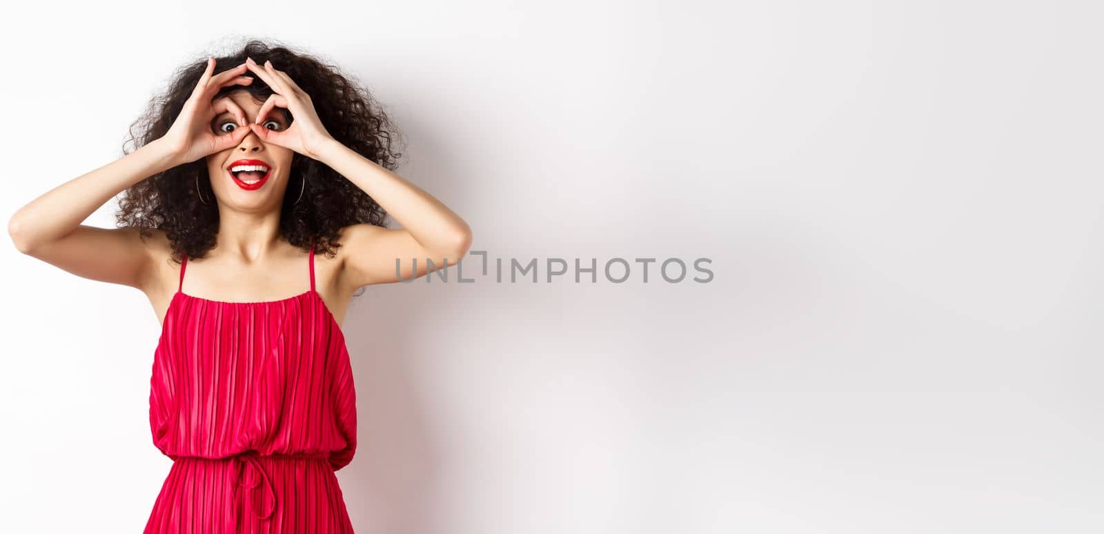 Impressed young woman seeing cool thing, look through hand binoculars and say wow, standing amazed at white background.