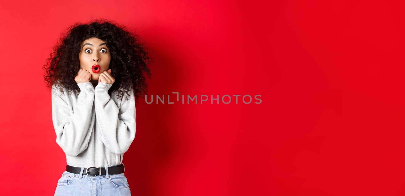 Surprised young woman looking with disbelief and amazement at camera, saying wow and standing in awe on red background.