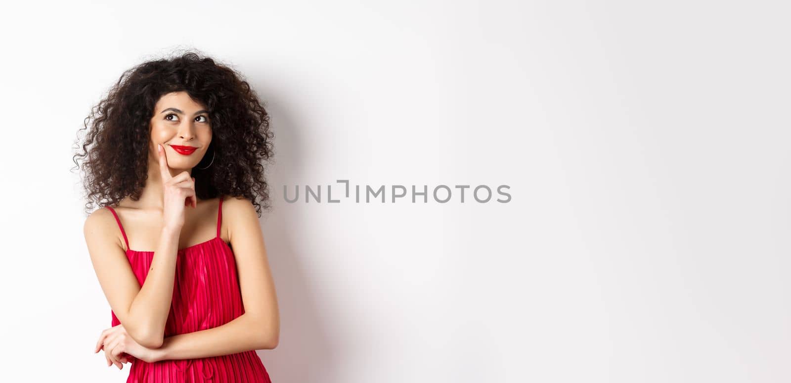 Image of elegant woman in red dress, looking pensive and smiling, thinking of romantic date, standing over white background.