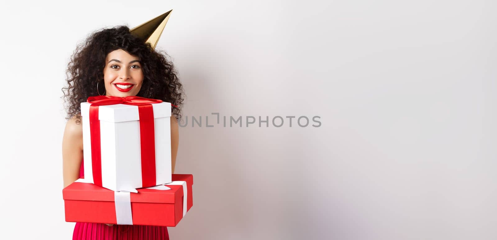 Holidays and celebration. Beautiful lady with curly hair, wearing party hat and holding big presents, smiling happy, receiving birthday gifts, standing on white background by Benzoix