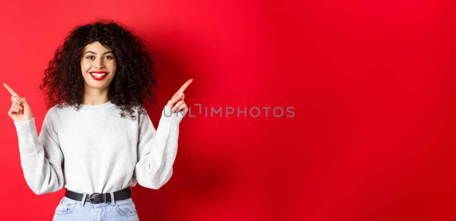 Attractive caucasian woman showing way, pointing fingers sideways at two promos, demonstrating variants and smiling, standing on red background.