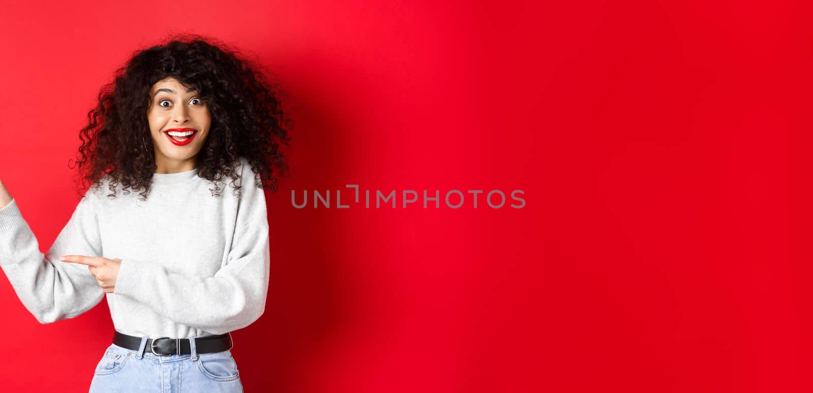 Happy woman with curly hair, smiling amused, pointing fingers left at logo and looking amazed, checking out special deal, standing on red background.