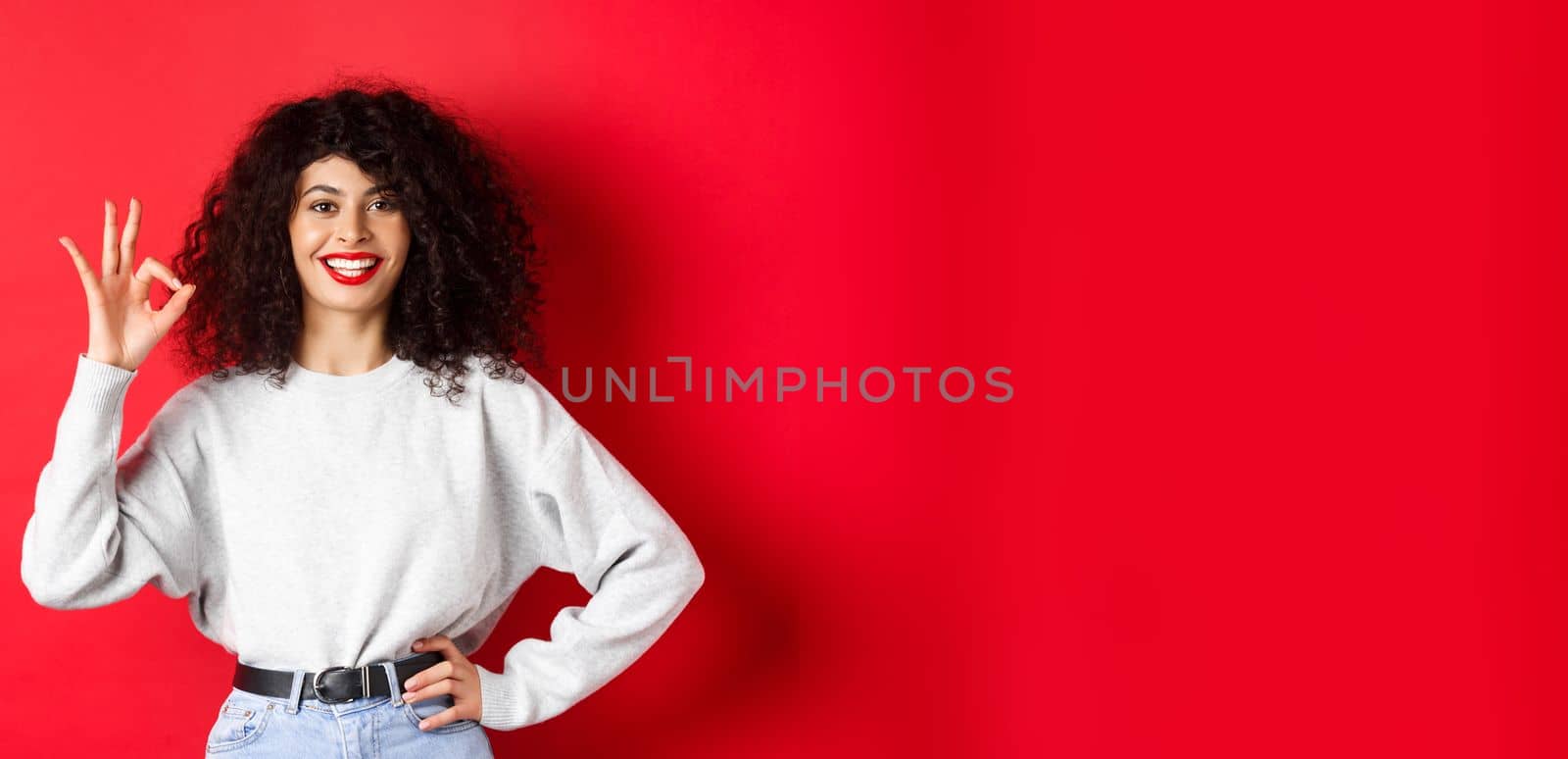 Confident smiling woman in casual clothes, showing OK sign to approve or like product, guarantee good quality, making a compliment, red background.