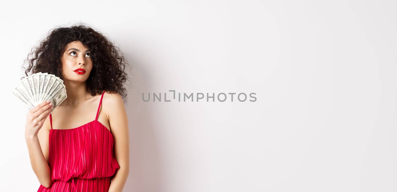 Elegant beautiful woman in red dress, waving with dollars and looking fabulous, standing on white background. Copy space