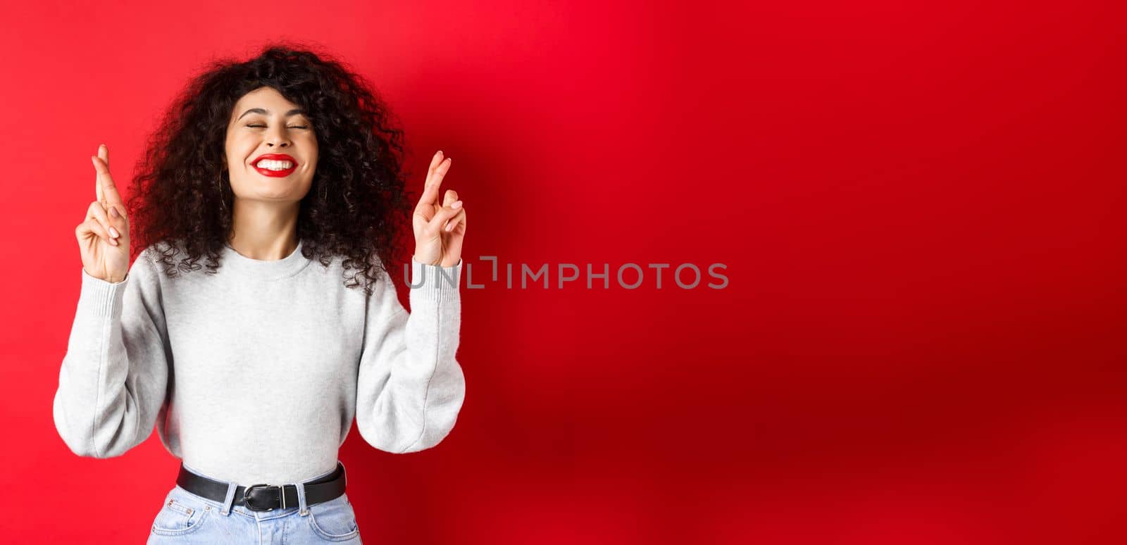 Hopeful and positive woman with red lips and curly hair, cross fingers for good luck and making wish, praying for dream come true, smiling excited, red background.