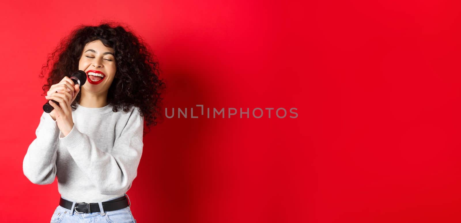 Hobbies and leisure concept. Happy woman singing song in microphone, having fun at karaoke with mic, standing on red background.