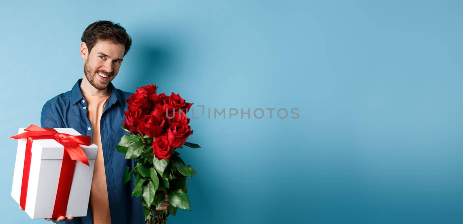 Love and Valentines day concept. Romantic smiling man giving you gift box and bouquet of flowers on date, standing over blue background.