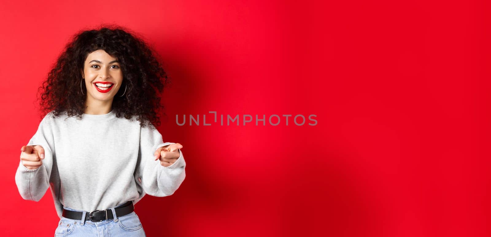 Cheerful woman with curly hair inviting you, recruiting newbies, pointing fingers at camera and smiling, praising good work, standing on red background.