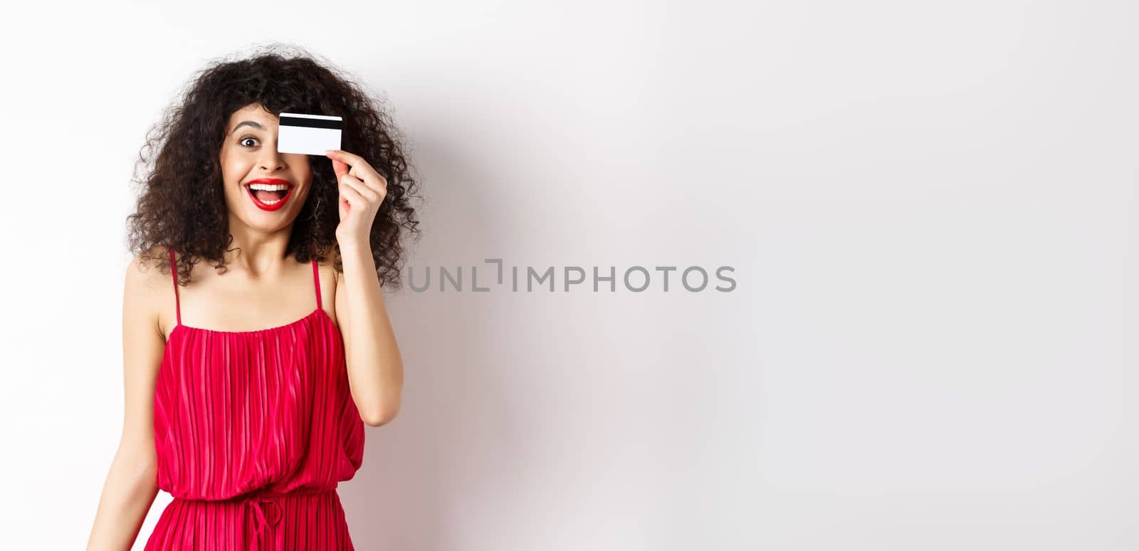 Shopping. Excited curly girl in red dress, showing plastic credit card and scream from joy, amazed with discounts and prices, standing against white background by Benzoix